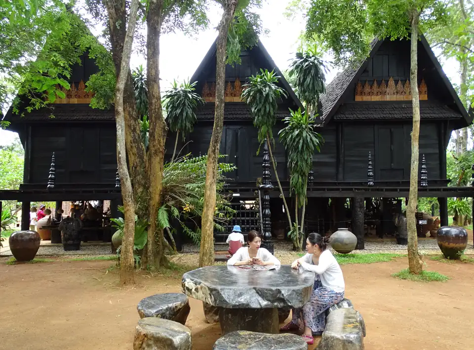 Black teak buildings on stilts with Lanna gables among trees, with stone furniture in front
