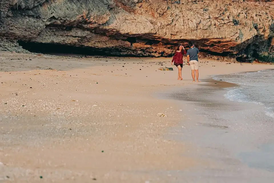 Two people alone on an empty beach beneath an overhanging cliff, one photographing the other
