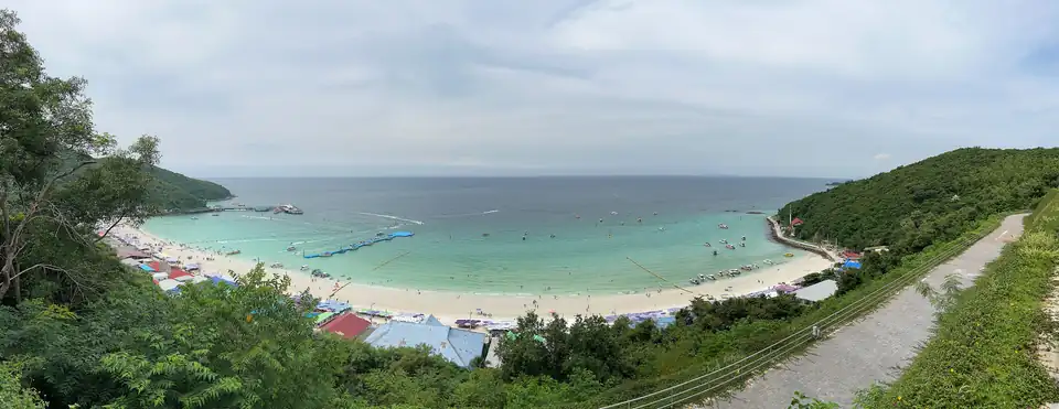 Wide view over Tawaen Beach on Koh Larn: a horseshoe bay with jetties, moored boats, sunloungers and a dense row of stalls behind the sand