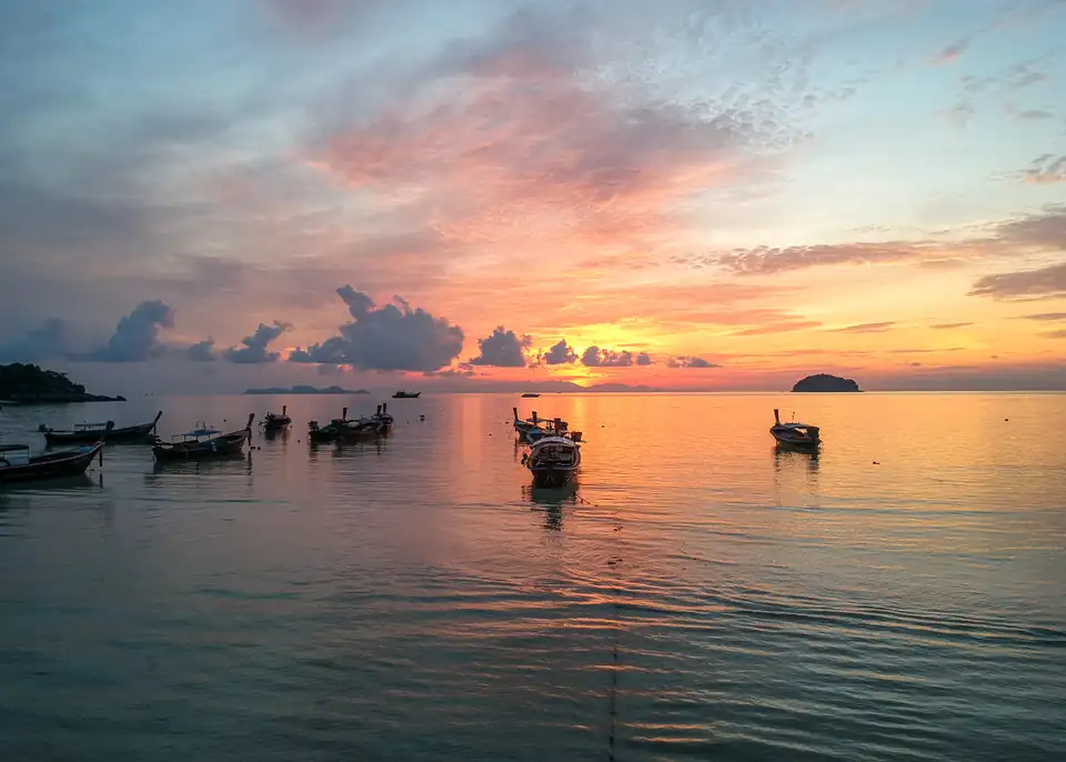 Long-tail boats moored on glassy water at dawn off Sunrise Beach, with pink and orange sky