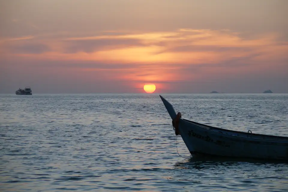 The bow of a long-tail boat silhouetted against a low orange sun over calm sea