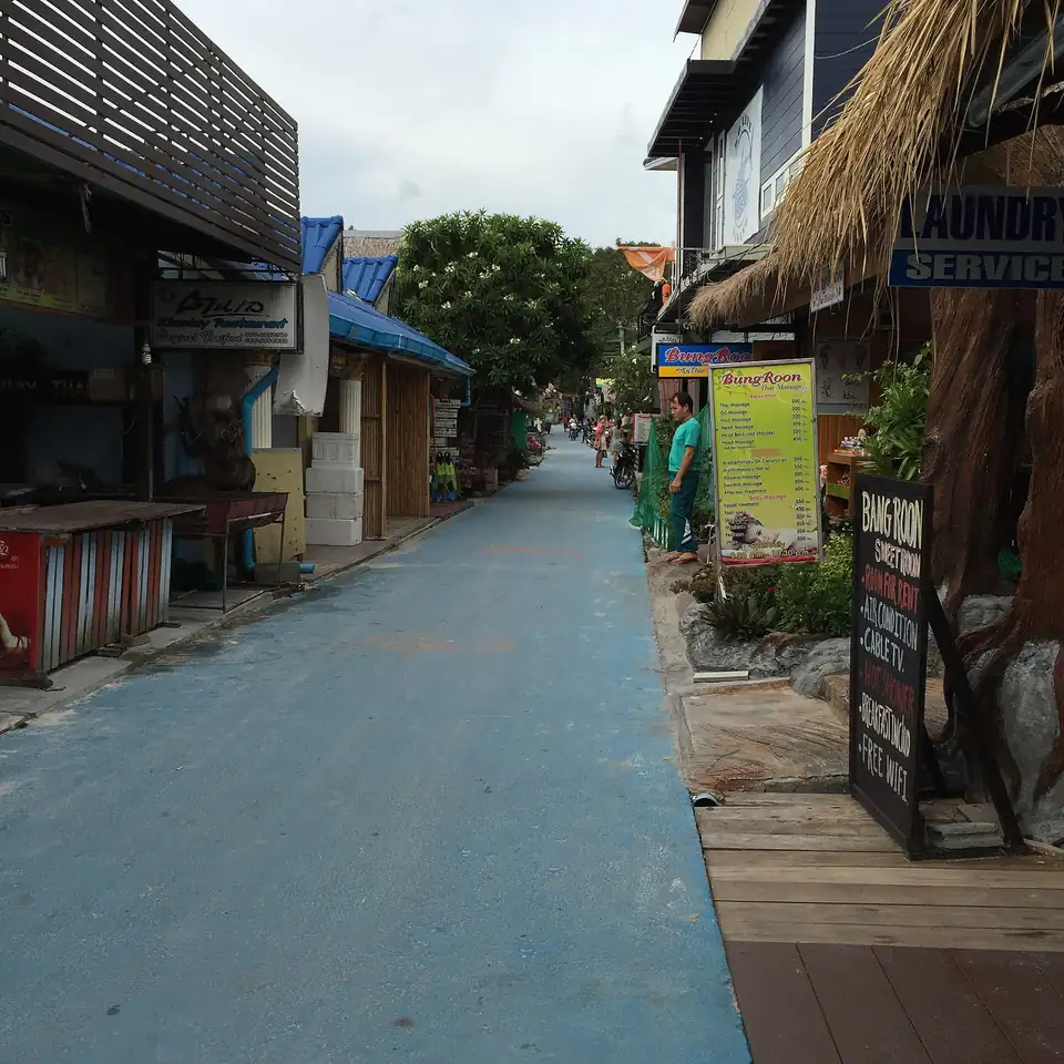 A narrow blue-painted concrete lane between low shopfronts, massage boards and a laundry sign