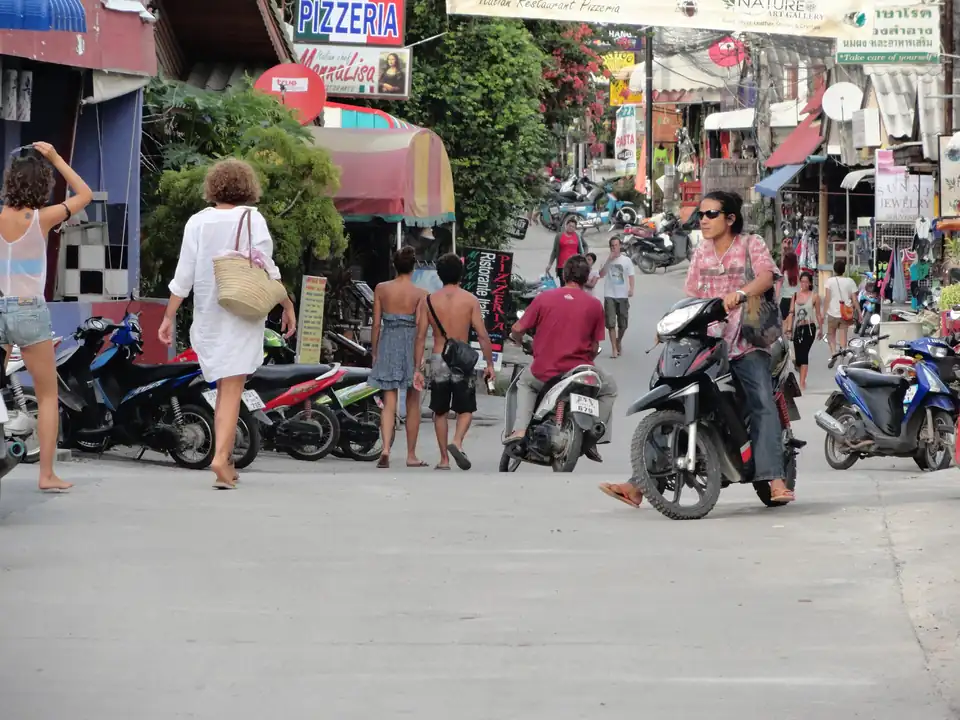 A busy street in Haad Rin lined with pizzerias, souvenir shops and parked scooters, with people walking through