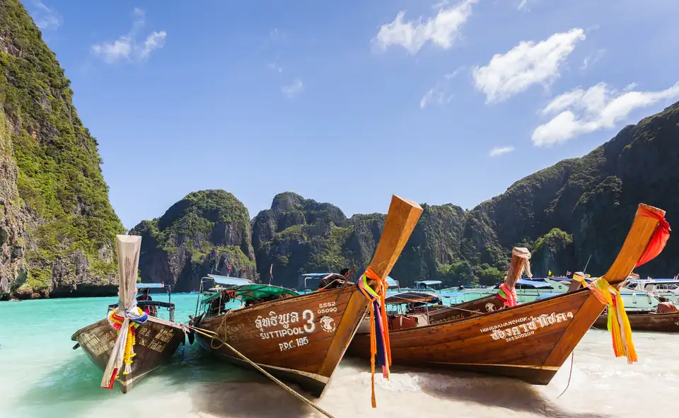 Long-tail boats drawn up on the sand in Maya Bay with limestone cliffs behind and a fleet of boats beyond