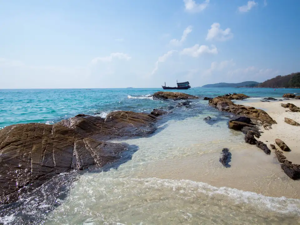 White sand, a low rock outcrop and clear turquoise water on Koh Samet, with a wooden boat offshore