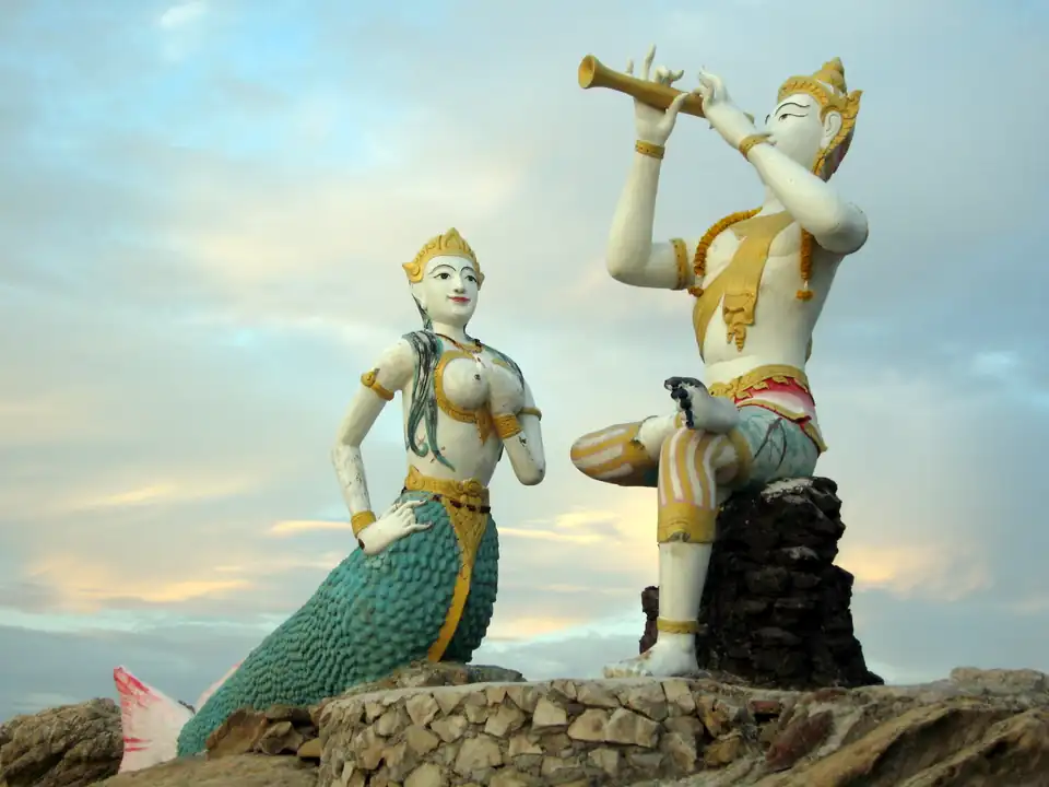 Two painted statues on a rock at dusk: a mermaid and a seated prince playing a flute