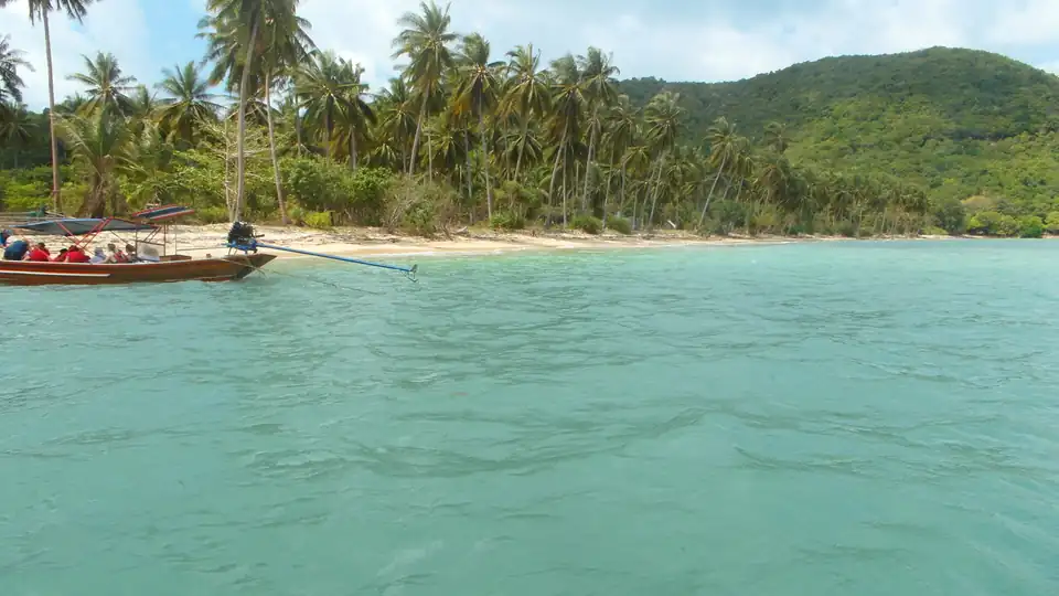 The wooded shore of Ko Tan seen from the water, with a longtail boat at anchor off a narrow beach