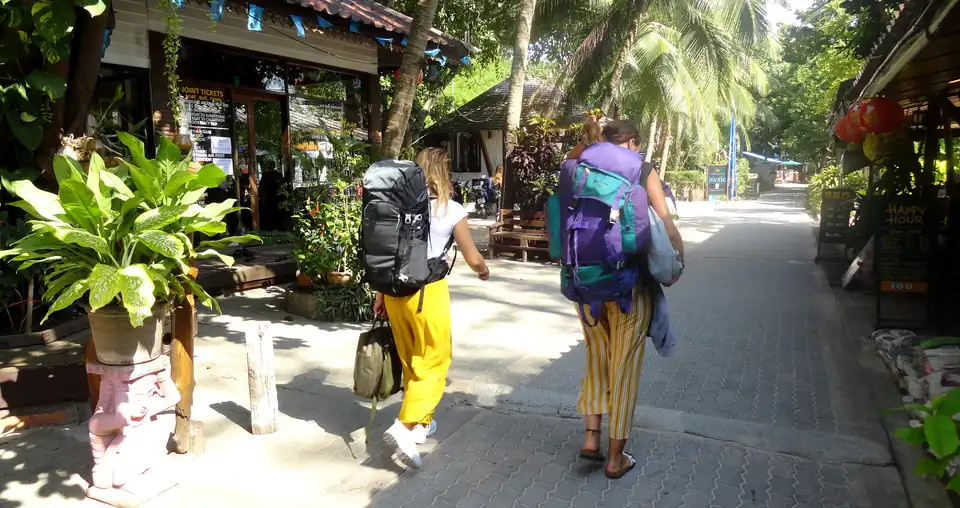 Two travellers with rucksacks walking a narrow shaded lane past a ticket agent, dive shop signs and a happy hour board