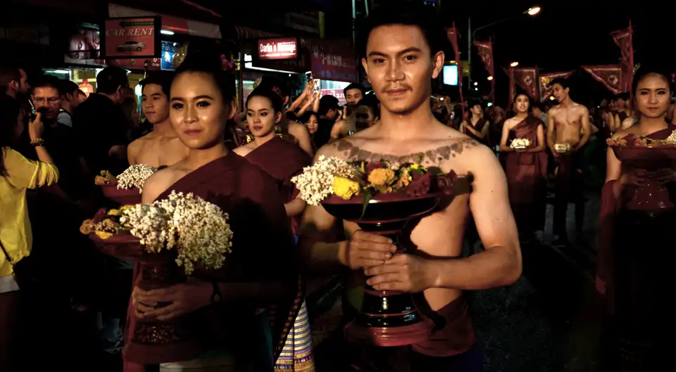 Participants in traditional Lanna dress carrying flower-filled offering bowls along a street at night