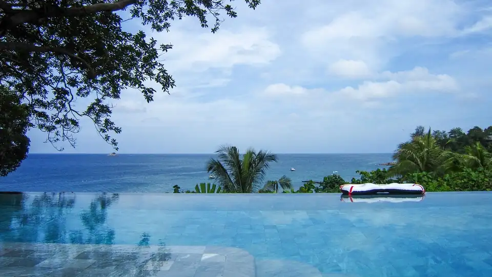 An infinity pool with the sea beyond its far edge, a tree leaning in from one side and a single rolled sunbed mattress left on the lip