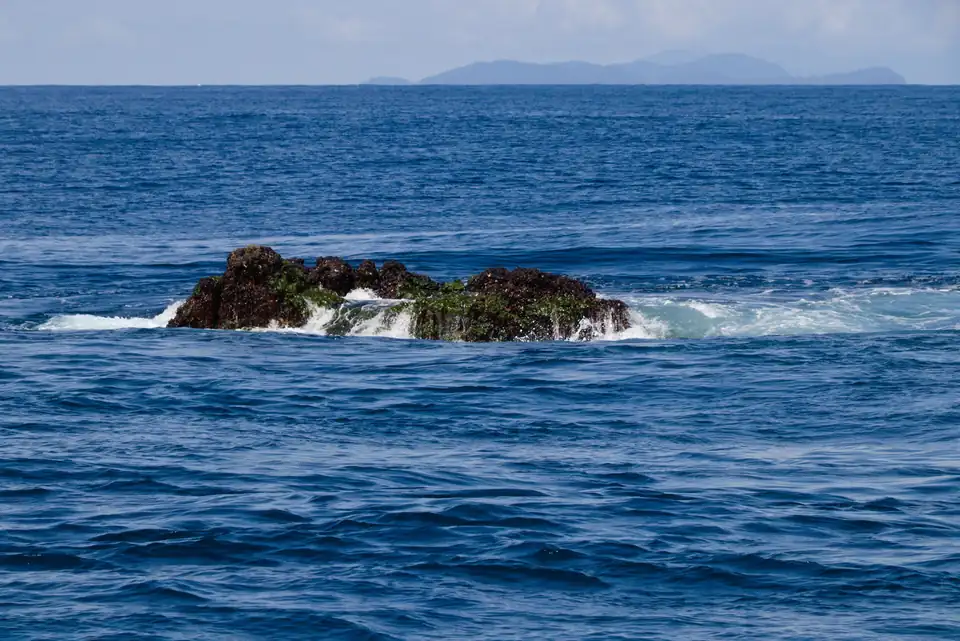 A small dark rock breaking the surface of open blue ocean with distant islands on the horizon